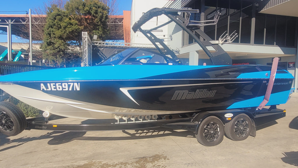 A blue and black Malibu brand speedboat on a dual-axle trailer, parked outdoors on a concrete surface with a building and trees in the background.