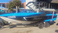 A blue and black Malibu brand speedboat on a dual-axle trailer, parked outdoors on a concrete surface with a building and trees in the background.