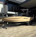 A beige and black MasterCraft ski and boat trailer is parked outside a dealership on a concrete lot under a clear blue sky.