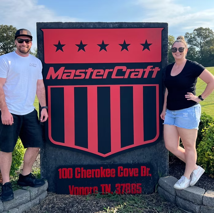 Two people standing outdoors on either side of a large MasterCraft sign. The sign is black and red with five black stars and the MasterCraft logo, plus the address '100 Cherokee Cove Dr.