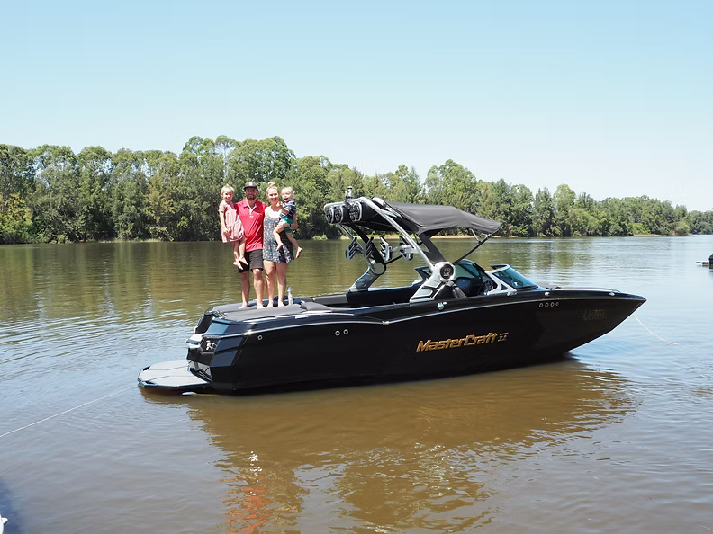 A black MasterCraft xstar wakeboat on a lake with trees in the background, with four people standing on the deck of the boat.