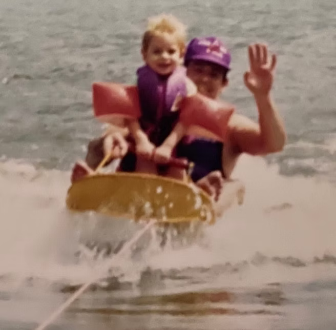 A person in a purple swim cap and dark swimsuit waterslide with a young child wearing a purple life jacket and pink floatation arms on a yellow tow board in the water.