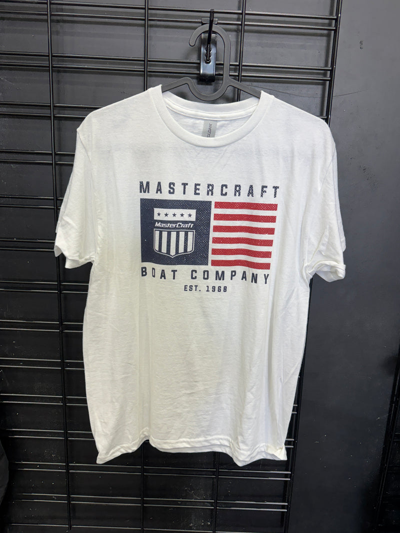 A white short-sleeve t-shirt with a vintage-style graphic print featuring a red and white striped flag, a navy blue shield, and the text 'MASTERCRAFT BOAT COMPANY EST. 1986', laid flat on a black wire rack.