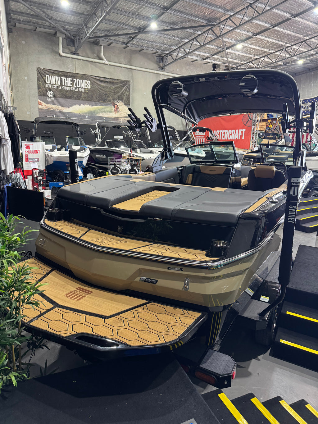 A beige and black motorboat with a hardtop and rear ramp, photographed inside a boat showroom. The boat features tan wood-look deck flooring and a black hull, with a T-handle tower and black seating.