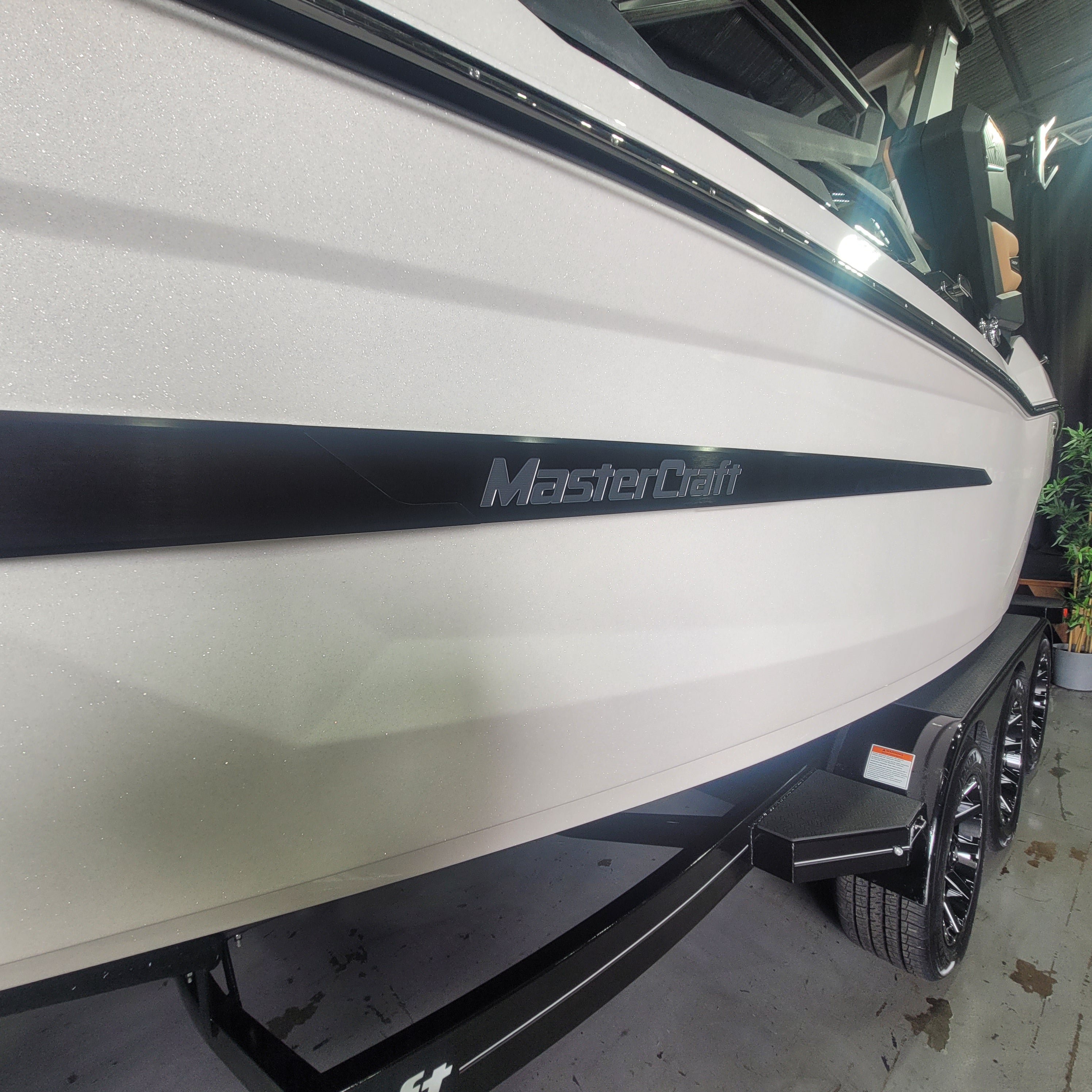 Close-up side view of a white MasterCraft boat on a trailer, showing the side stripe, brand name, hull, wheels, and part of the cabin in an indoor showroom or garage setting.