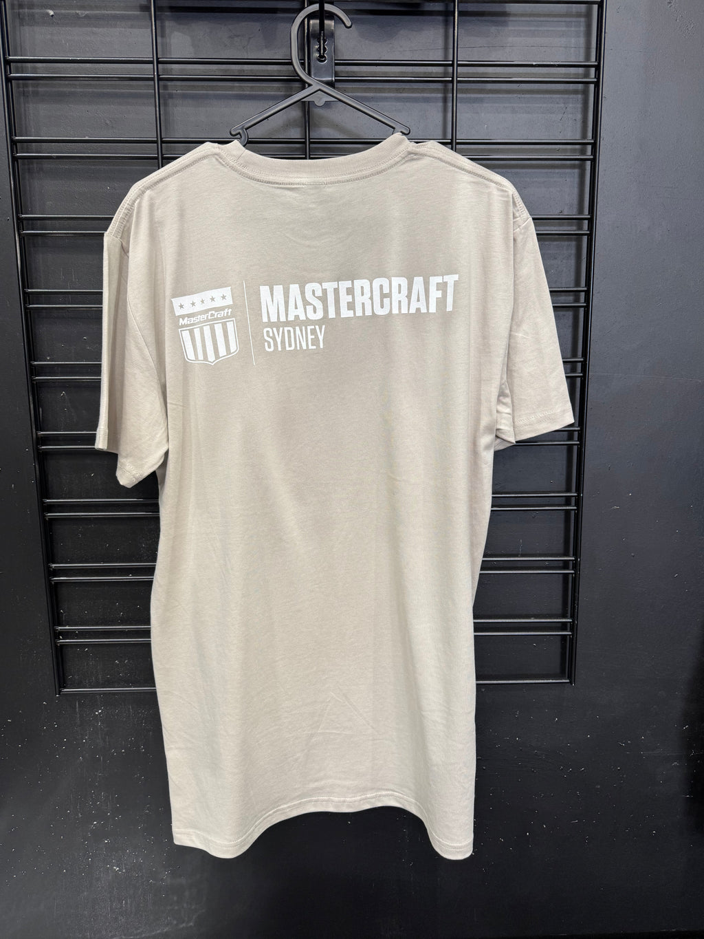 A light beige crewneck t-shirt hanging on a black hanger against a black metal grid wall. The back of the shirt features a white graphic print with the text 'MASTERCRAFT SYDNEY' and a shield logo.