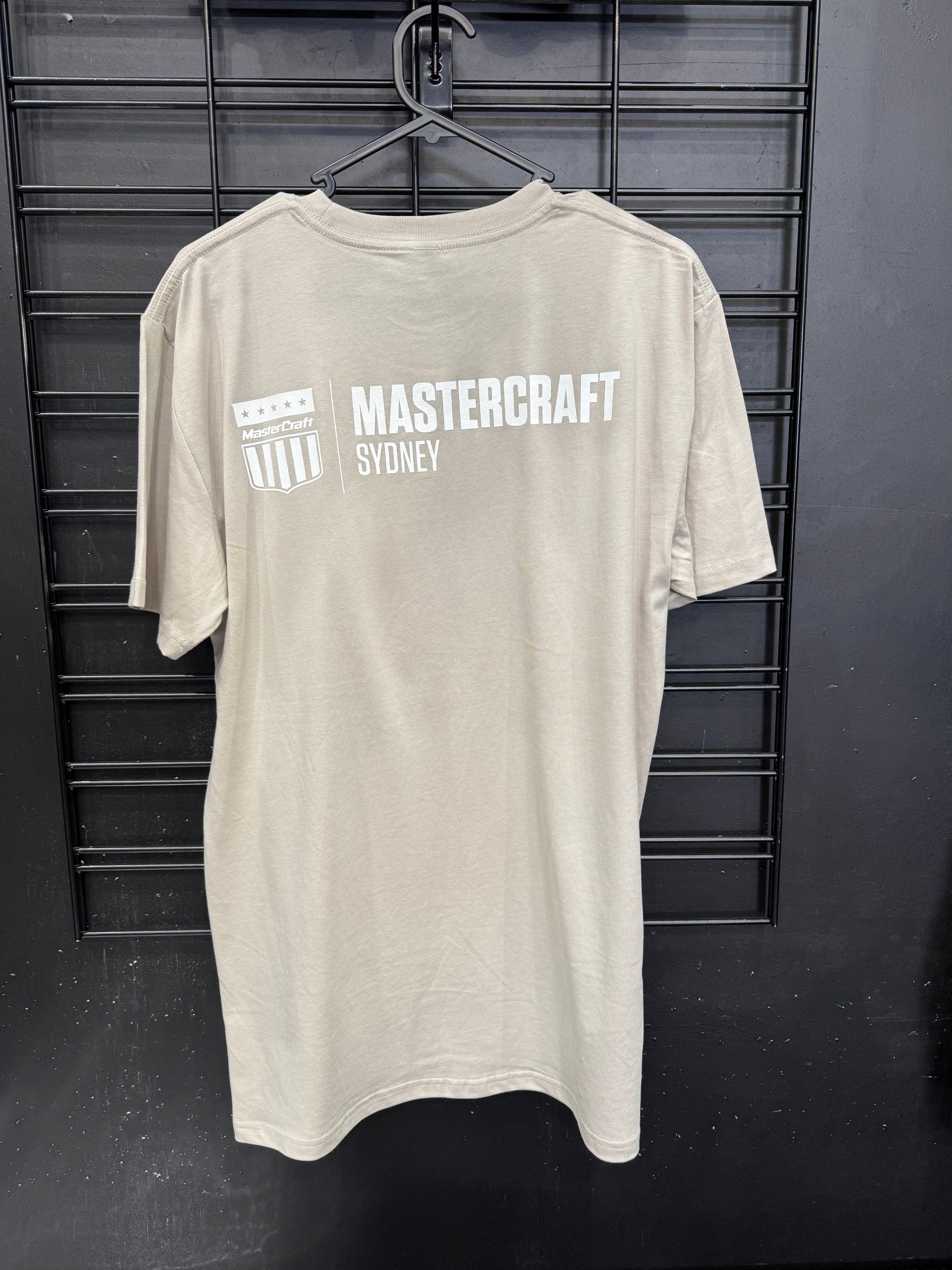A light beige crewneck t-shirt hanging on a black hanger against a black metal grid wall. The back of the shirt features a white graphic print with the text 'MASTERCRAFT SYDNEY' and a shield logo.