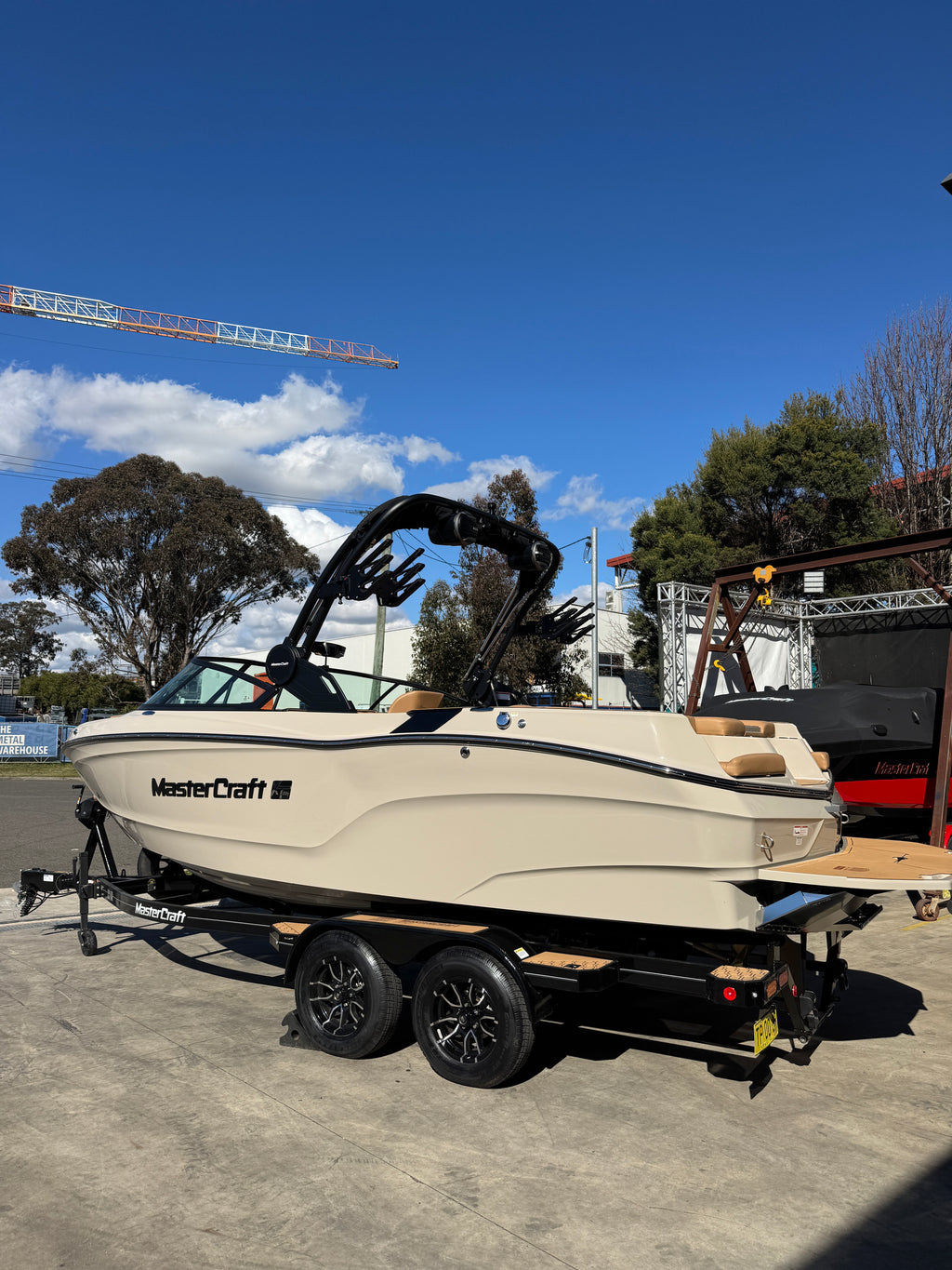 A MasterCraft wakeboat in cream and black, mounted on a black dual-axle trailer, photographed outdoors on a concrete surface under a blue sky with trees and a crane in the background.