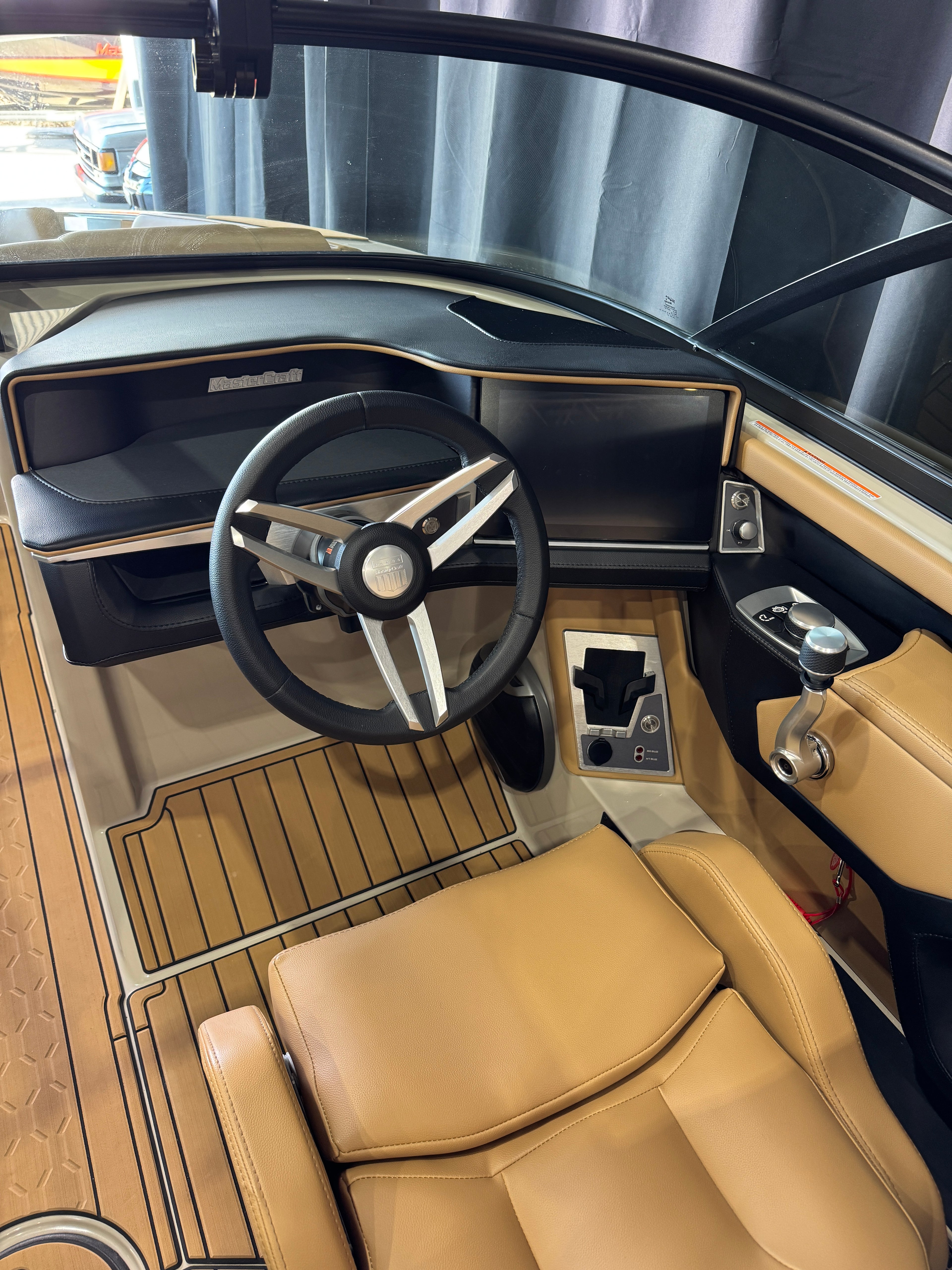 The helm station of a luxury MasterCraft XT22T Boat featuring tan leather seats, a black and silver three-spoke steering wheel, a curved dashboard with wood trim, and wood-look floor mats.