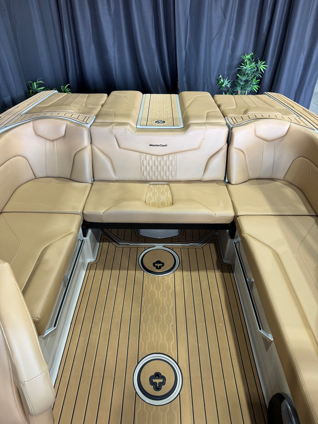 Beige leather U-shaped MasterCraft XT22T boat seating with a central console table and a wood-look floor, photographed indoors against dark blue curtains.