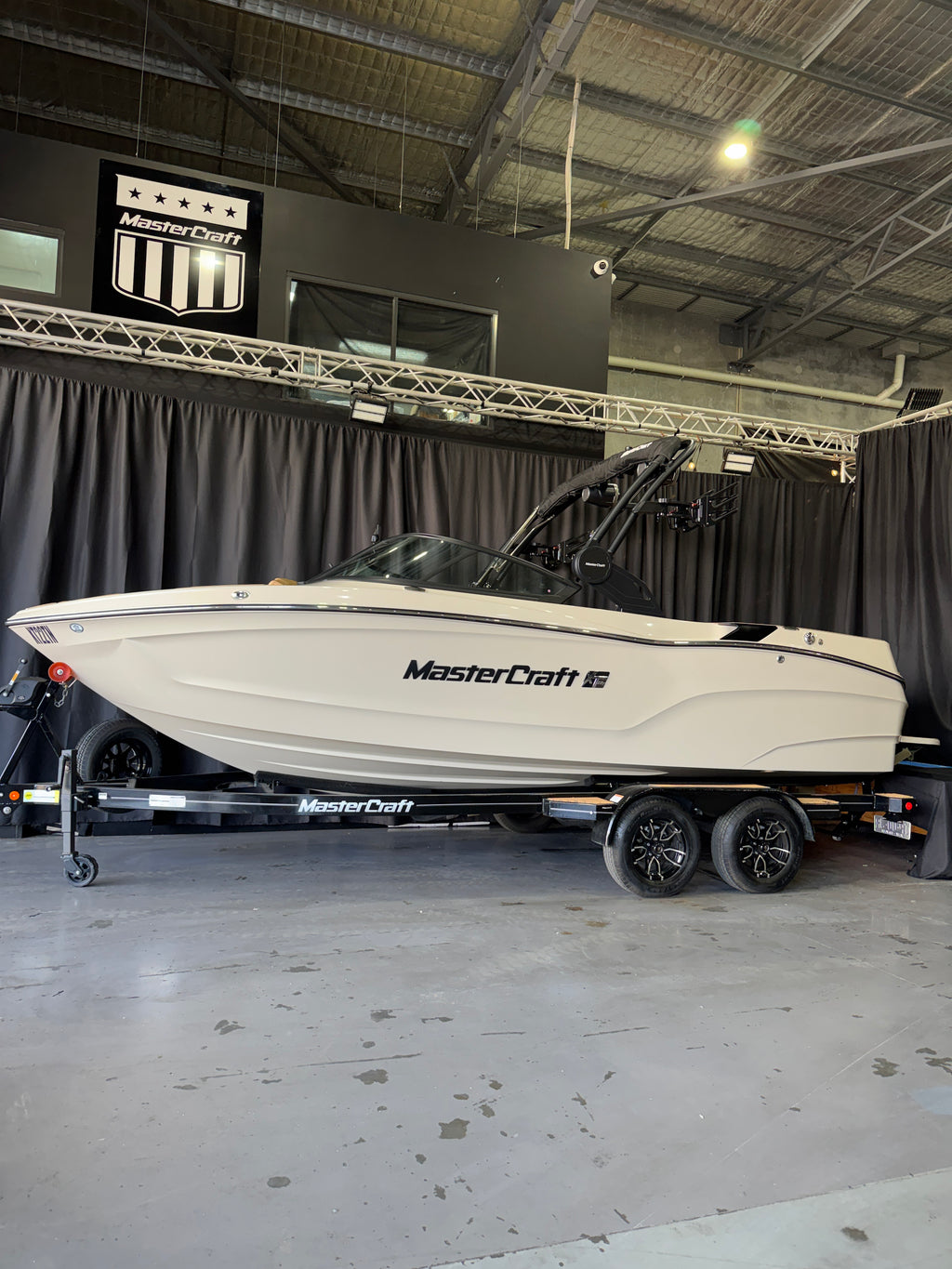 A white MasterCraft wakeboat on a black dual-axle trailer, photographed inside a warehouse or showroom with black curtains and a metal ceiling visible in the background.