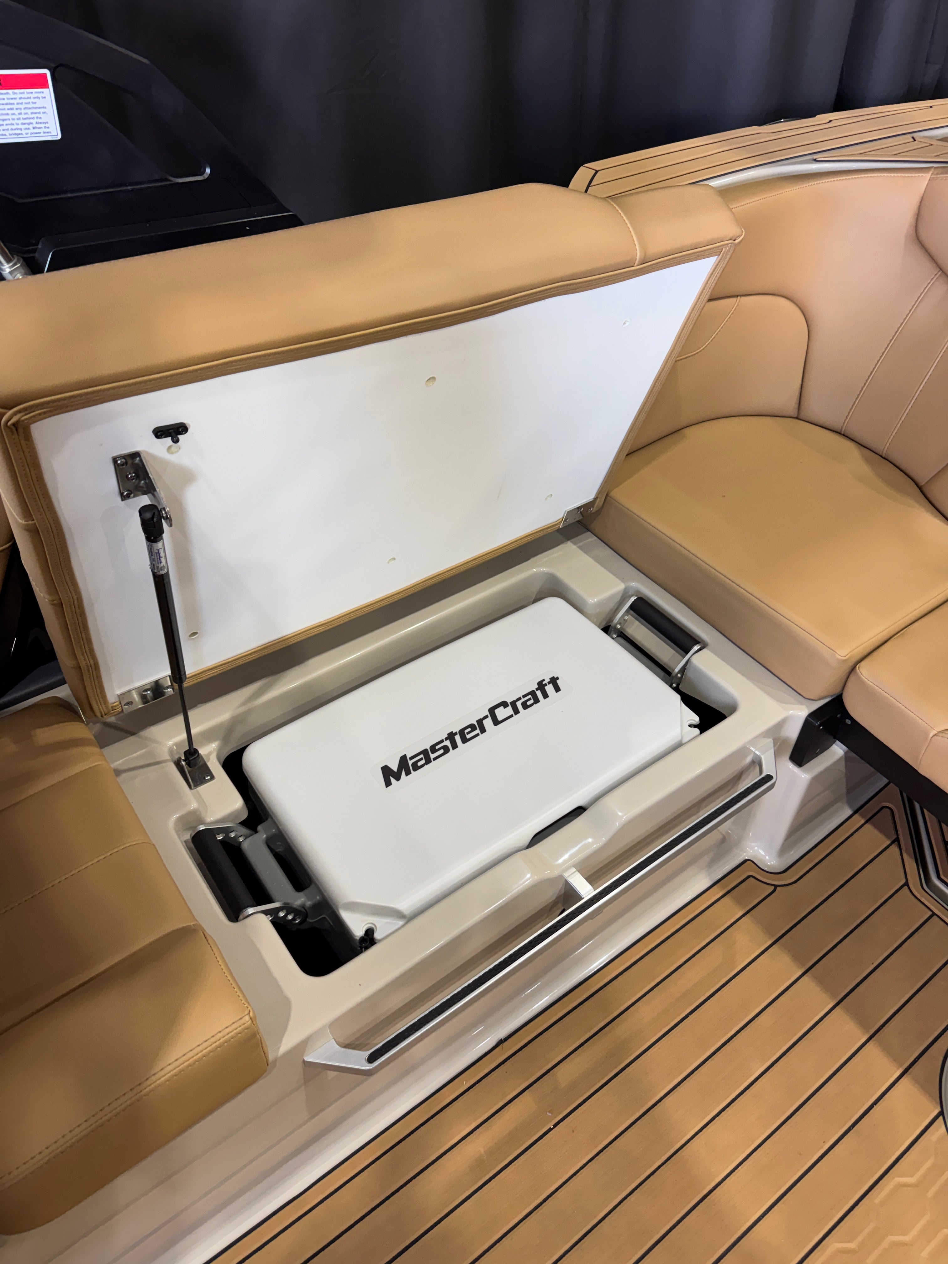 A white MasterCraft cooler or storage box placed inside an open beige storage compartment in a boat cabin, with tan upholstered seating and wood-look flooring visible around it.