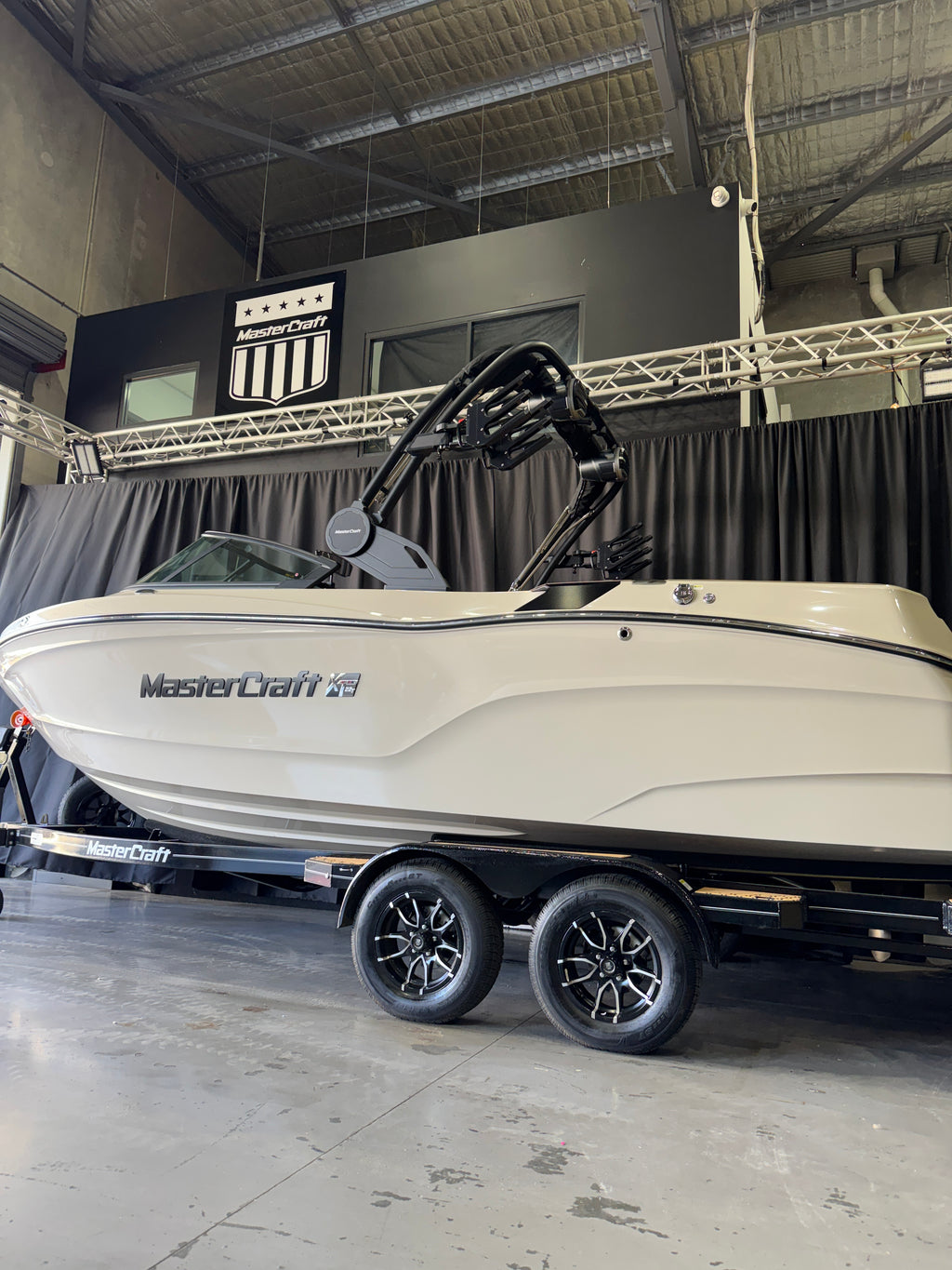 A white MasterCraft 28 boat on a black dual-axle trailer, photographed inside a warehouse or showroom with a black curtain backdrop and high industrial ceiling.
