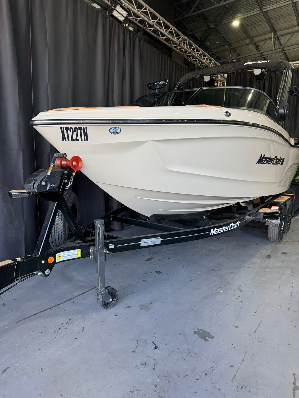 A STONE MasterCraft XT22T boat on a black dual-axle trailer inside a warehouse or showroom with a gray concrete floor and black curtain backdrop.