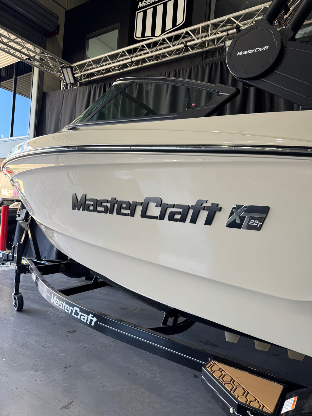 A white MasterCraft XT22T boat on a black trailer inside a showroom or warehouse. The MasterCraft branding is clearly visible on the side of the boat and the trailer frame.