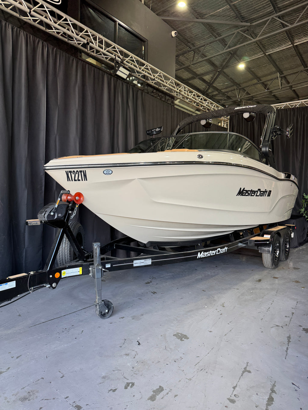 A MasterCraft XT22T boat on a black trailer inside a warehouse or showroom with dark curtains and a concrete floor.