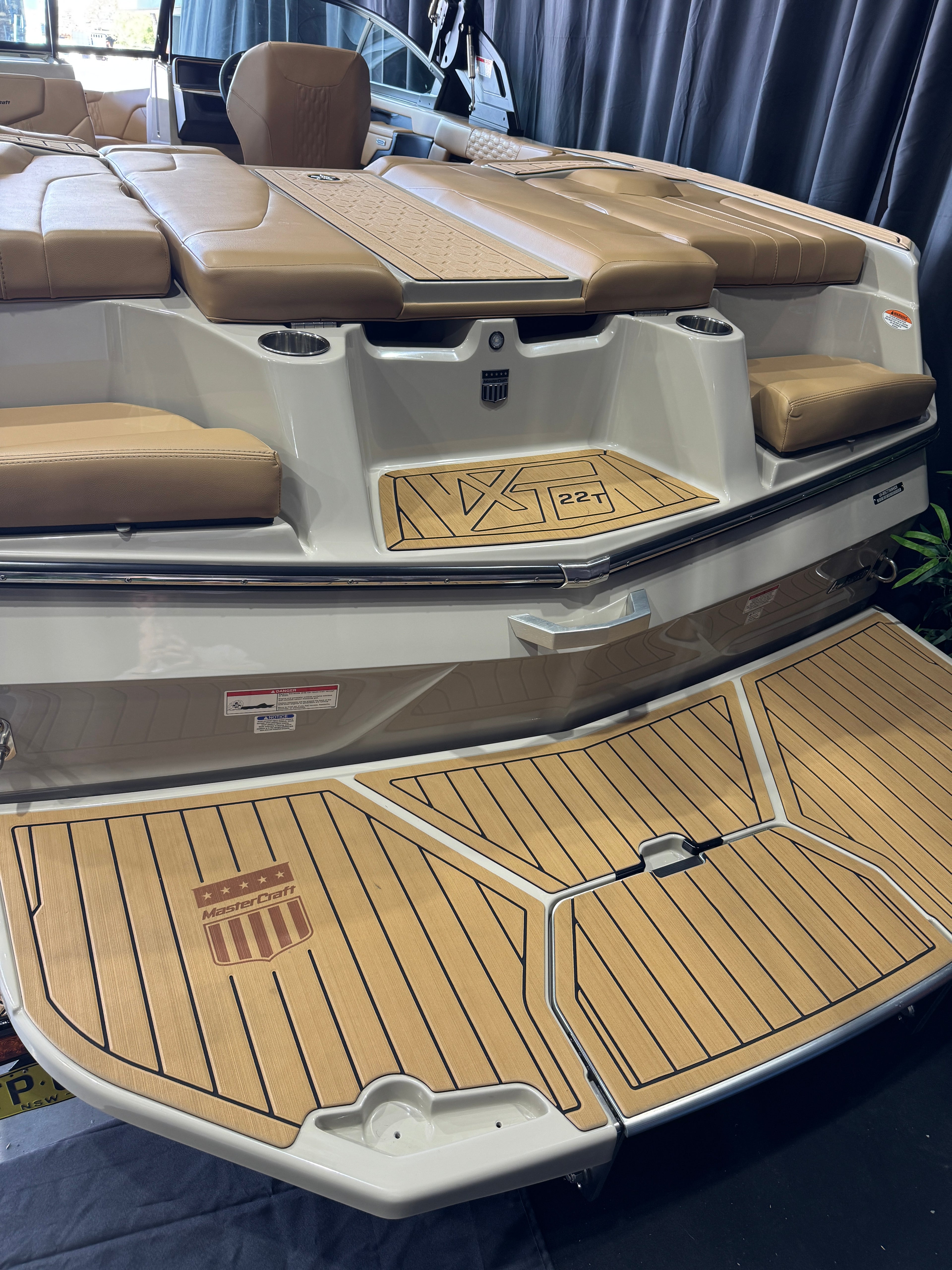 Close-up of the stern deck area of a MasterCraft XT22T boat with tan and beige upholstered seating, tan wood-look deck flooring, and cream-colored hull panels.