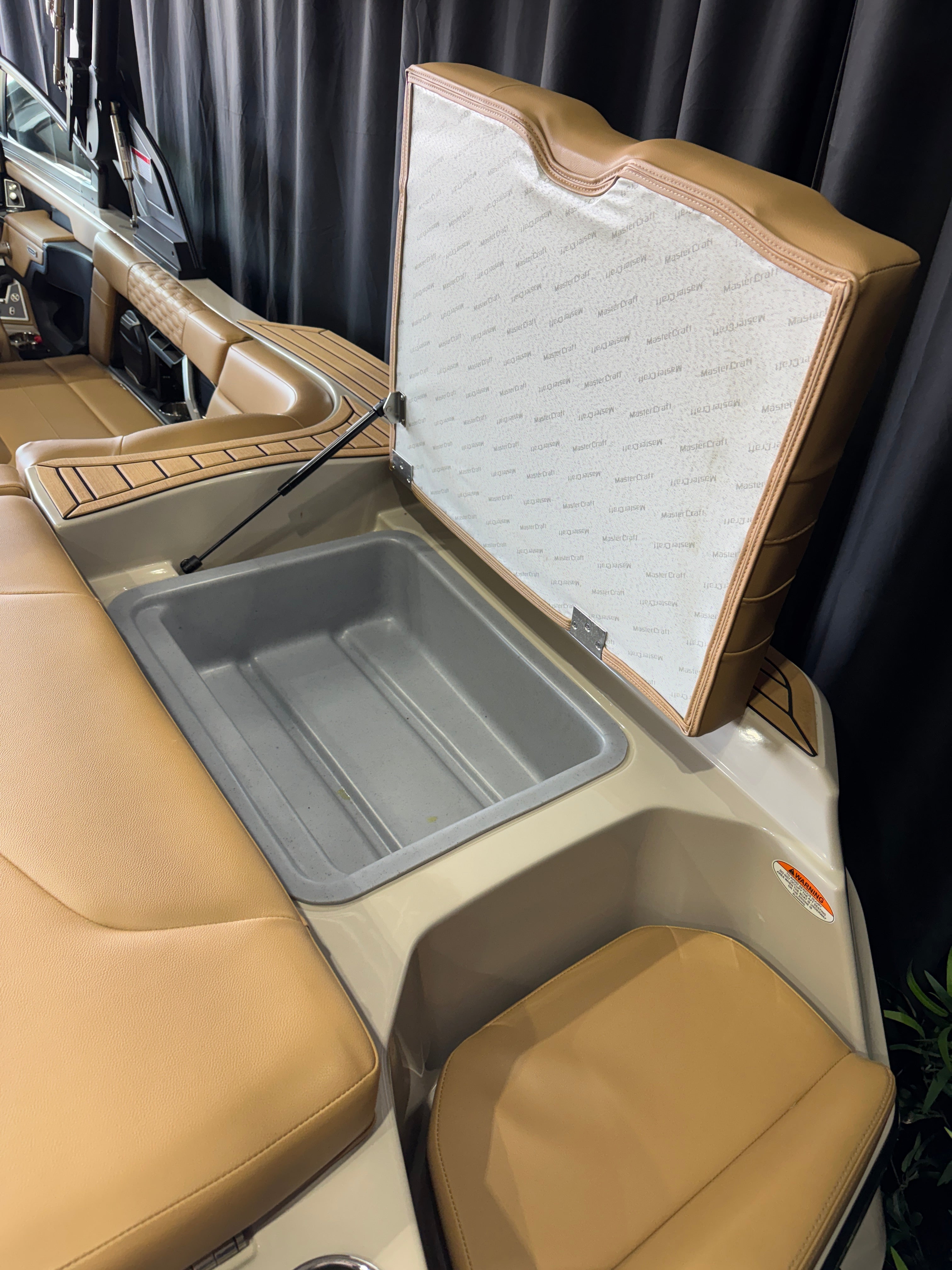 A close-up of a MasterCraft XT22T boat storage compartment with a tan leather seat, a gray plastic storage bin, and a hinged cover that is open to show a white textured interior lining.