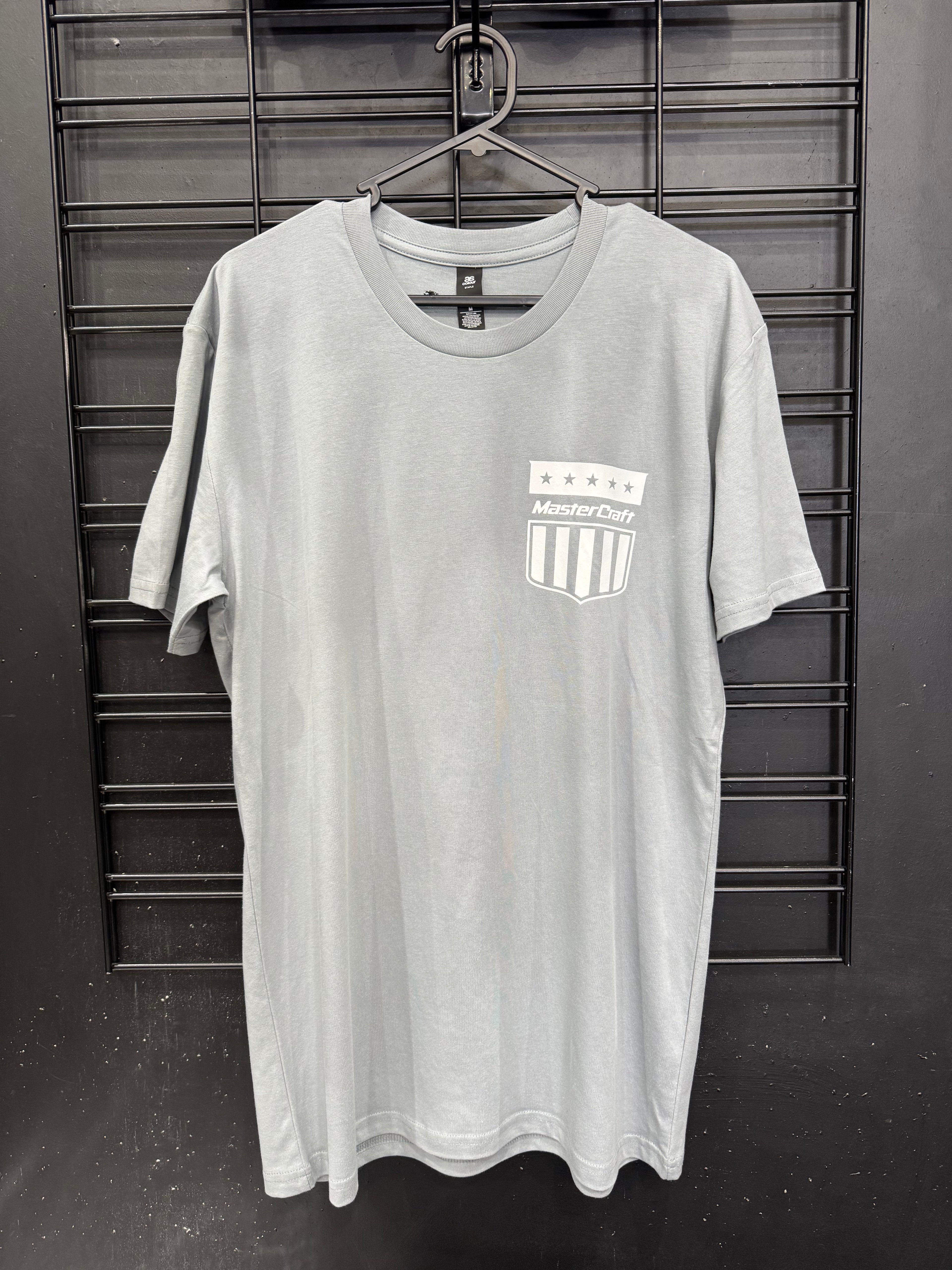 A light gray short-sleeve t-shirt hanging on a black hanger against a black metal grid wall. The shirt features a white and black shield crest on the left chest with the text 'MasterChill' and five stars above it.