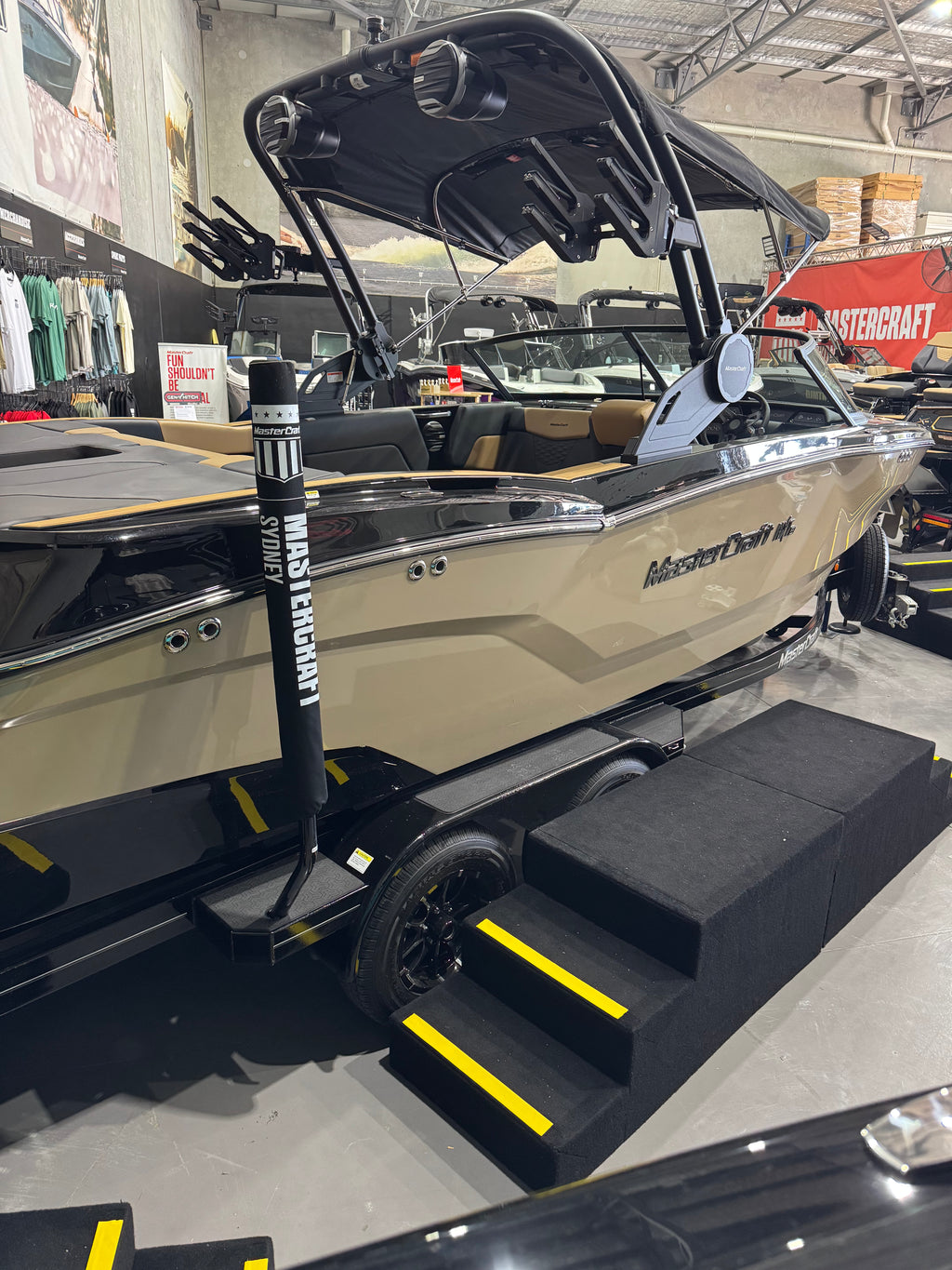 A beige and black MasterCraft wakeboat on a trailer inside a showroom, with a black bimini top, black steps, and yellow safety stripes.