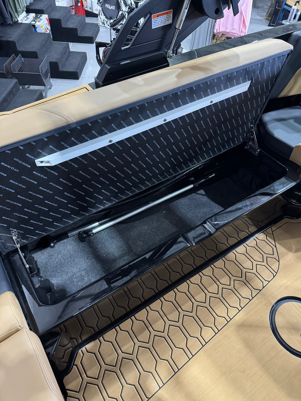 A close-up of a boat storage compartment with the seat back lifted, showing a black diamond-plate textured panel, a silver support bar, and a black interior. The surrounding area includes tan upholstery, a patterned floor mat