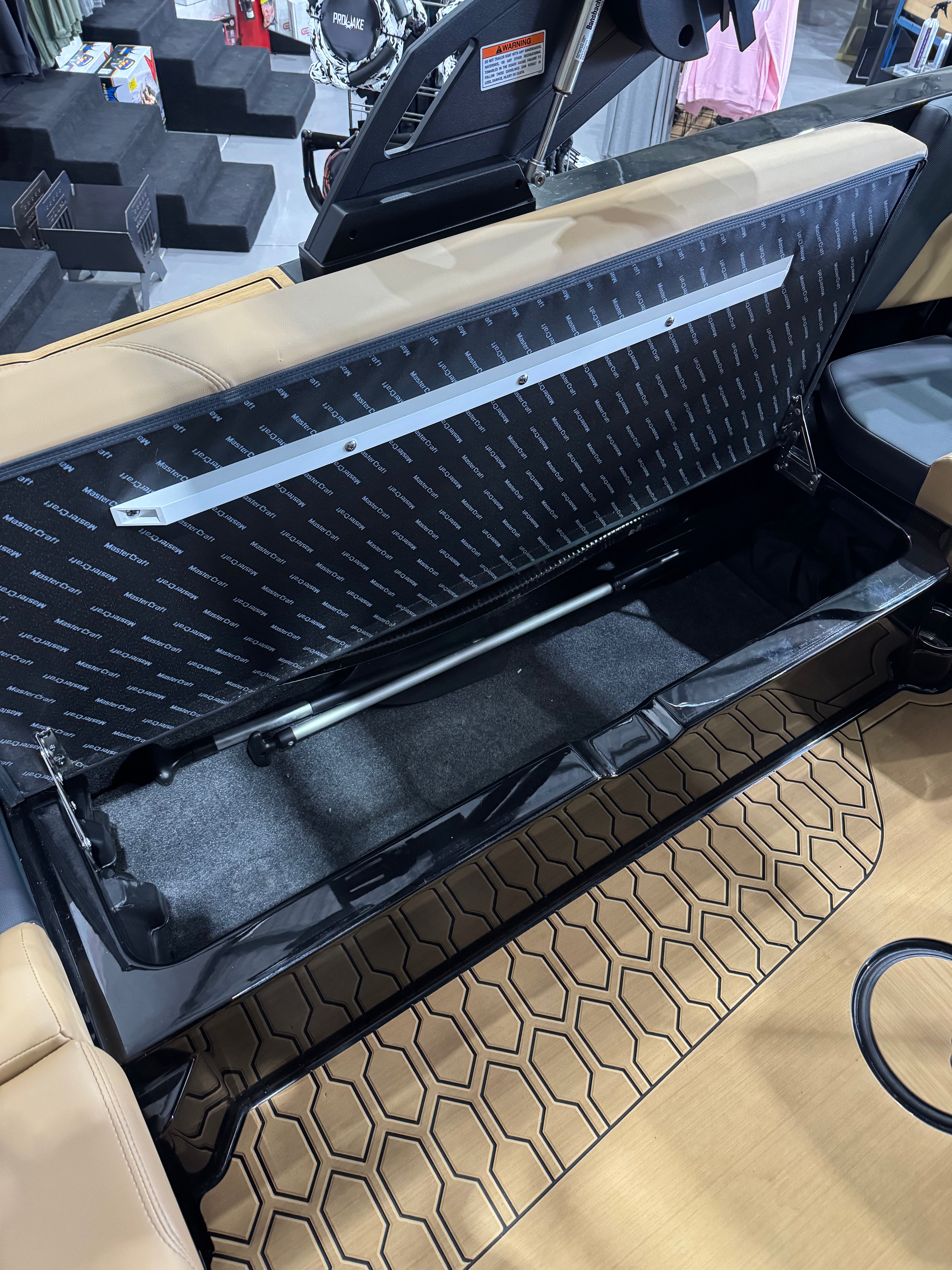 A close-up of a boat storage compartment with the seat back lifted, showing a black diamond-plate textured panel, a silver support bar, and a black interior. The surrounding area includes tan upholstery, a patterned floor mat