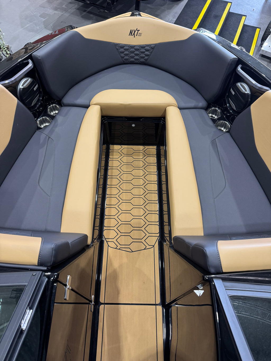 Close-up of the interior deck and seating of a boat with tan and dark gray upholstery, a wood-look floor, and black railings. The wordmark 'NXT' is visible on the backrest.