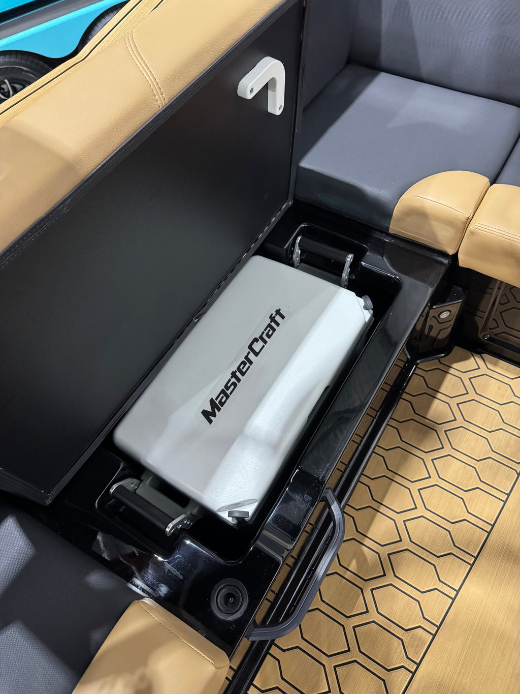 A white MasterCraft cooler or storage box placed inside a black storage compartment of a boat interior, with tan leather seating and a patterned floor visible around it.