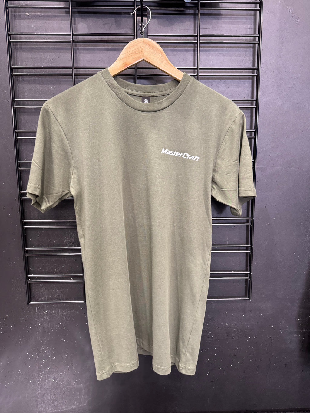 An olive green short-sleeve t-shirt with a white 'MasterCraft' logo printed on the left chest, hanging on a wooden hanger against a black wire grid wall.
