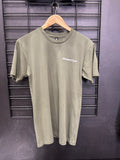 An olive green short-sleeve t-shirt with a white 'MasterCraft' logo printed on the left chest, hanging on a wooden hanger against a black wire grid wall.