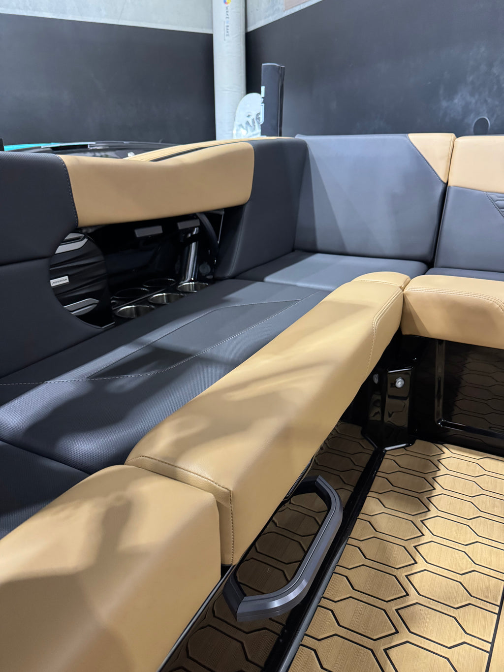 A close-up of a modern U-shaped boat or vehicle seating area with tan and dark gray upholstered seats, black structural framing, and a patterned tan floor mat.