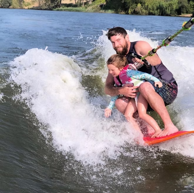A man and a child wakeboarding on a river, with the man holding the child close while they glide through white water. The man wears a black life vest and patterned swim trunks, and the child wears a pink and blue rash guard.