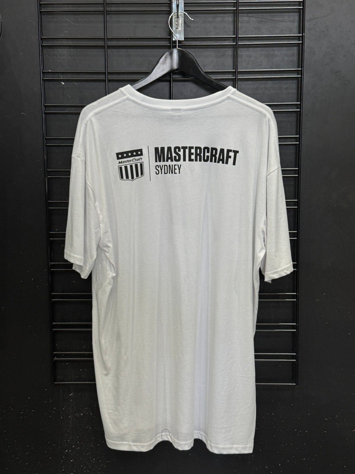 A white short-sleeve t-shirt hanging on a black hanger against a black slatted wall. The front of the shirt features a black graphic print on the left chest with a shield logo and the text 'MASTERCRAFT SYDNEY'.
