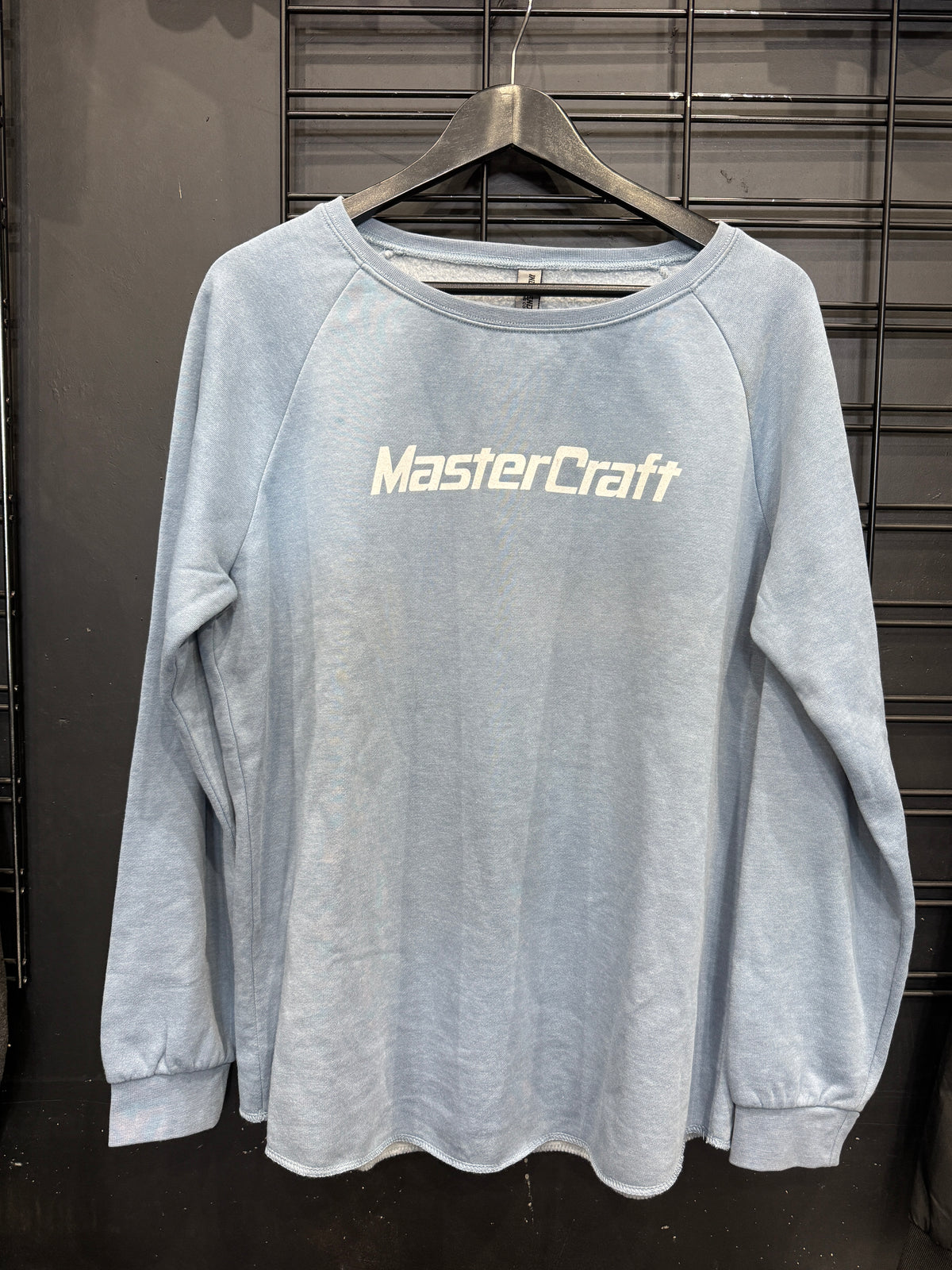 A light blue long-sleeve raglan sweatshirt with 'MasterCraft' printed in white across the chest, hanging on a black hanger against a dark metal grid background.