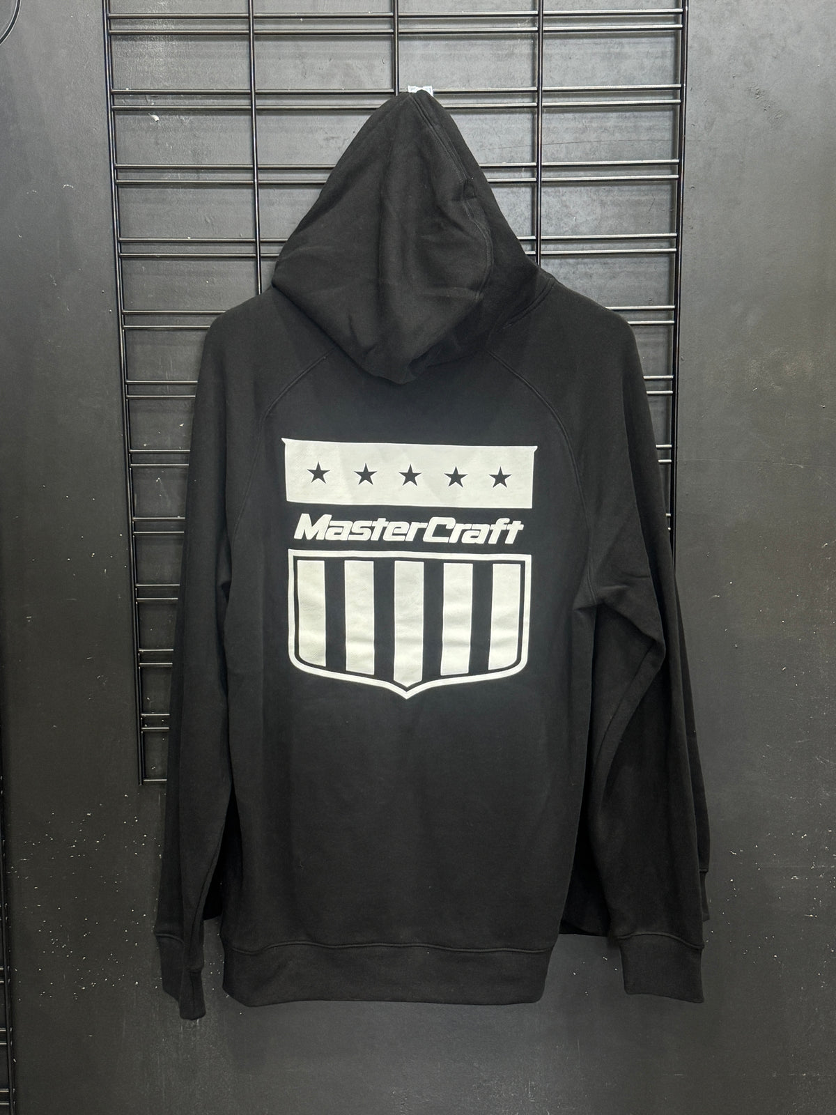 A black pullover hoodie with a white and gray MasterCraft shield logo and stars printed on the chest, laid flat on a dark metal wire rack.
