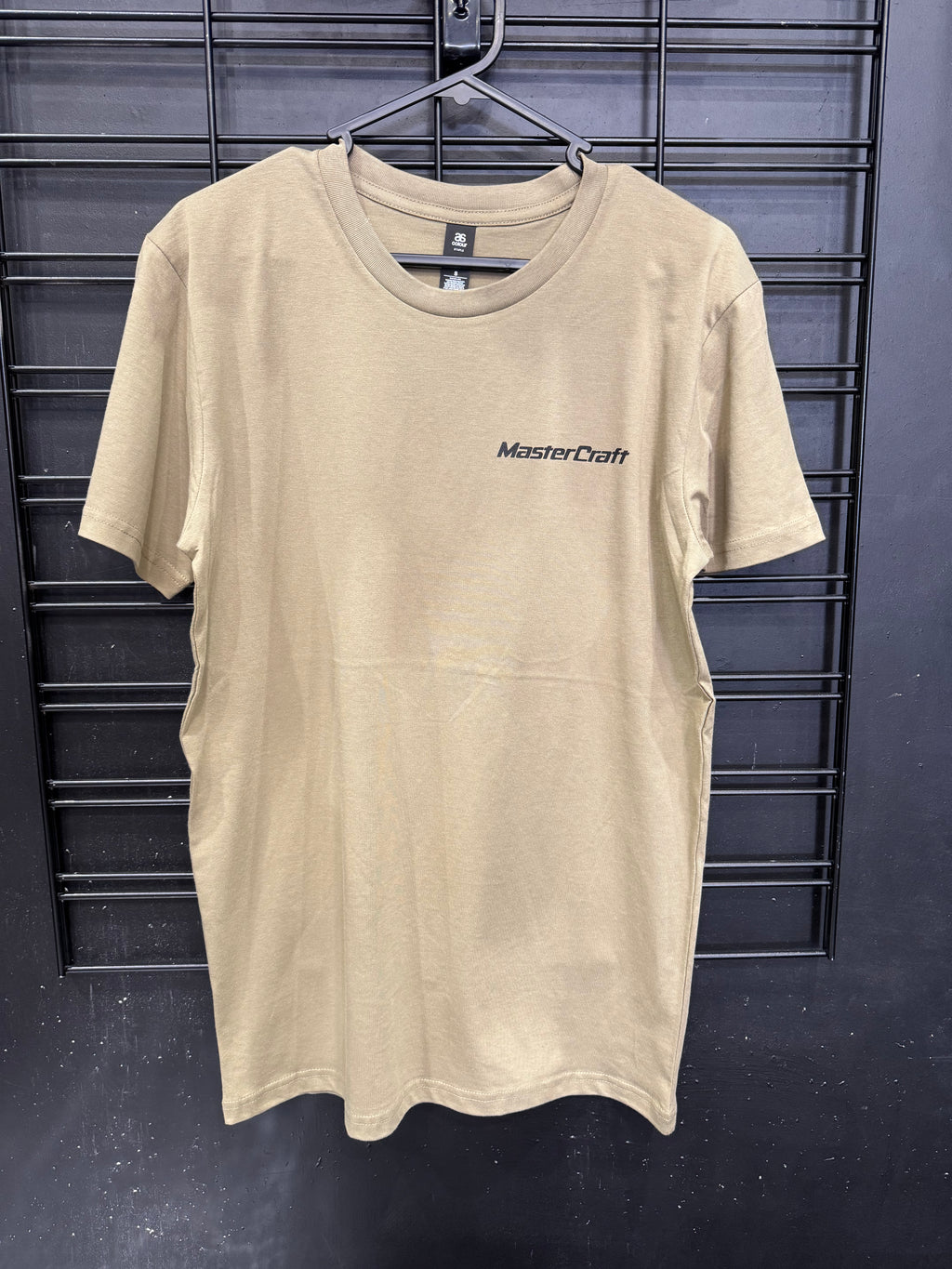 A beige short-sleeve crewneck t-shirt with 'MasterCraft' printed in black on the left chest, hanging on a black wire rack against a dark gray wall.