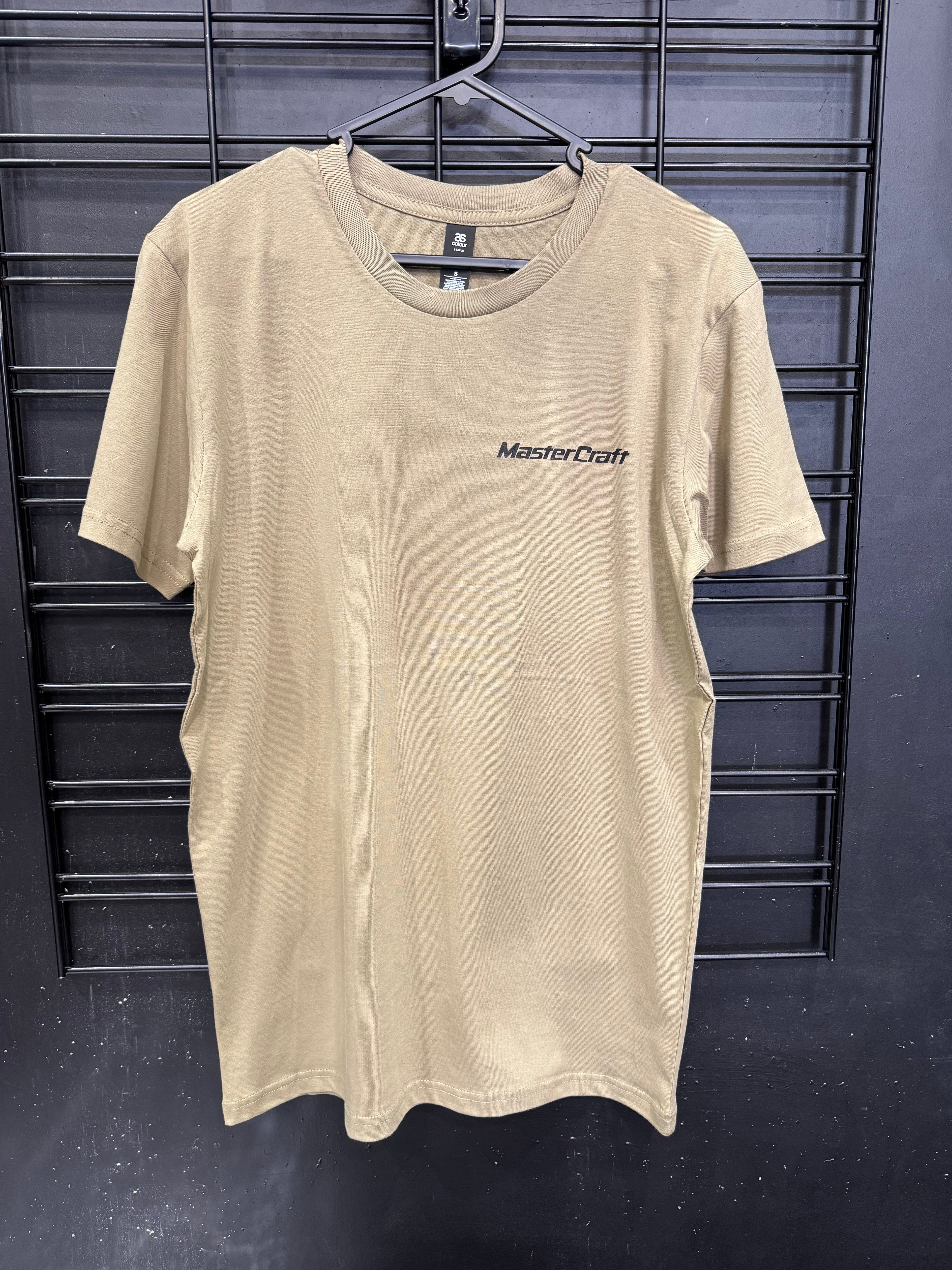 A beige short-sleeve crewneck t-shirt with 'MasterCraft' printed in black on the left chest, hanging on a black wire rack against a dark gray wall.