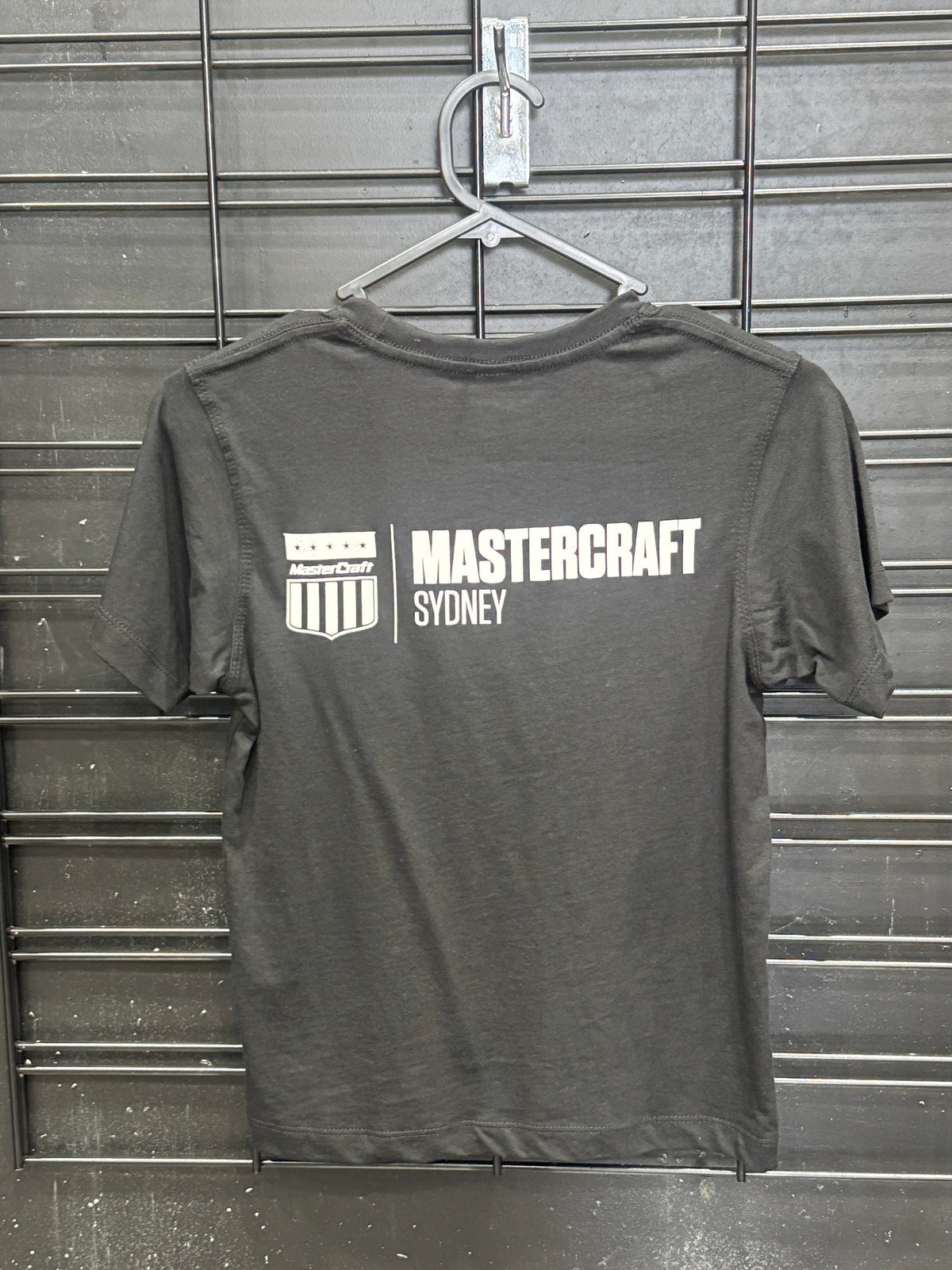 A dark gray heather t-shirt hanging on a metal hanger against a gray slatted wall. The back of the shirt features a white graphic print with the text 'MASTERCRAFT SYDNEY' and a shield logo.
