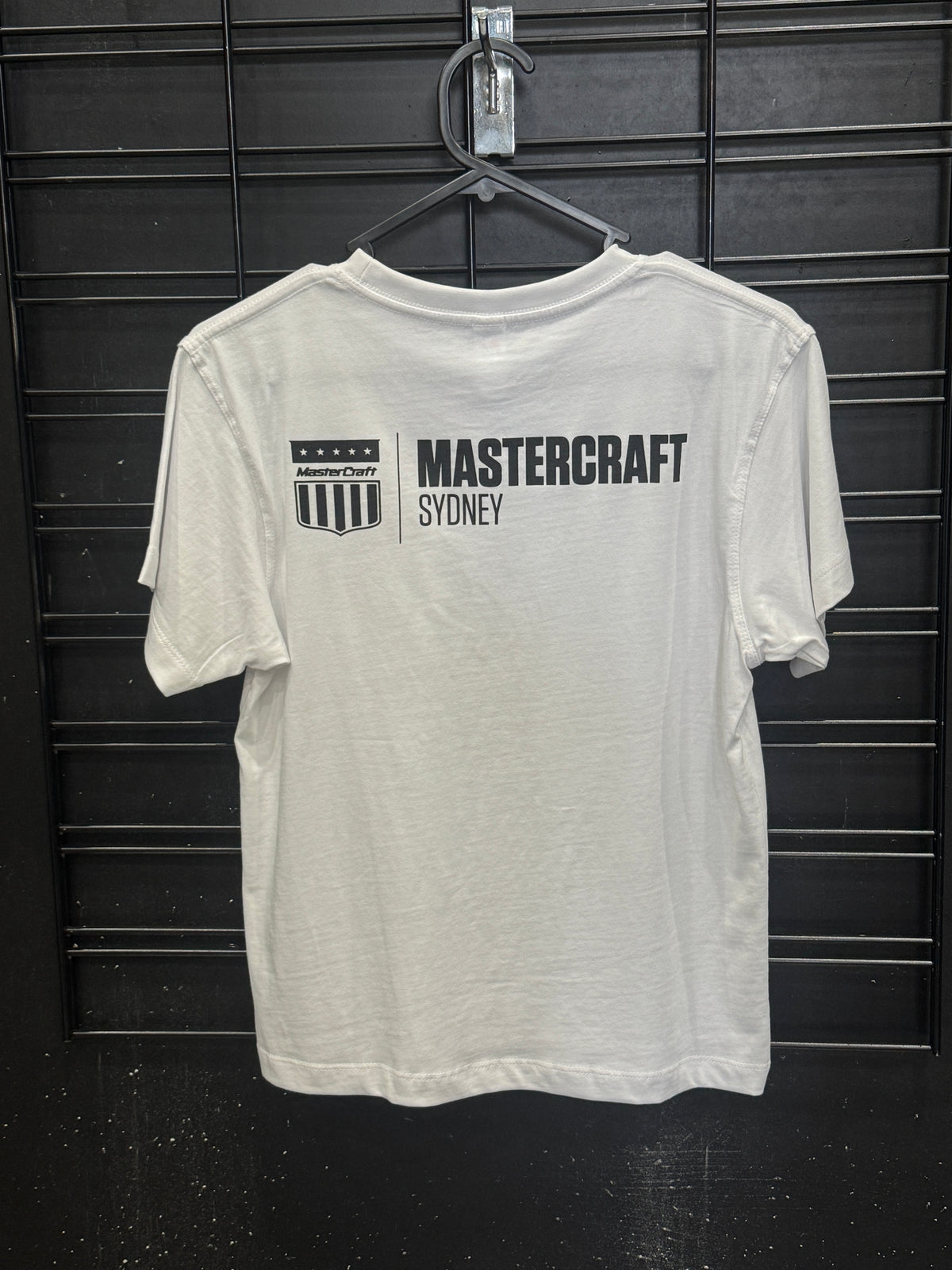 A white short-sleeve t-shirt with a black vertical graphic reading 'MASTERCRAFT SYDNEY' and a shield logo, displayed on a black wire hanger against a dark textured background.