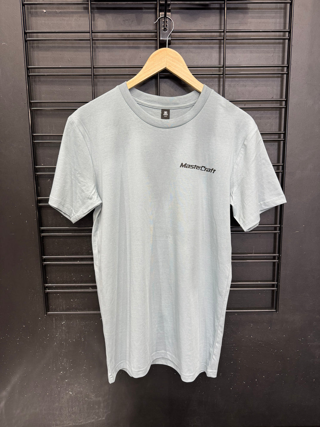 A light gray crewneck t-shirt with a small black 'MasterDuty' logo on the left chest, hanging on a wooden hanger against a black metal grid wall.