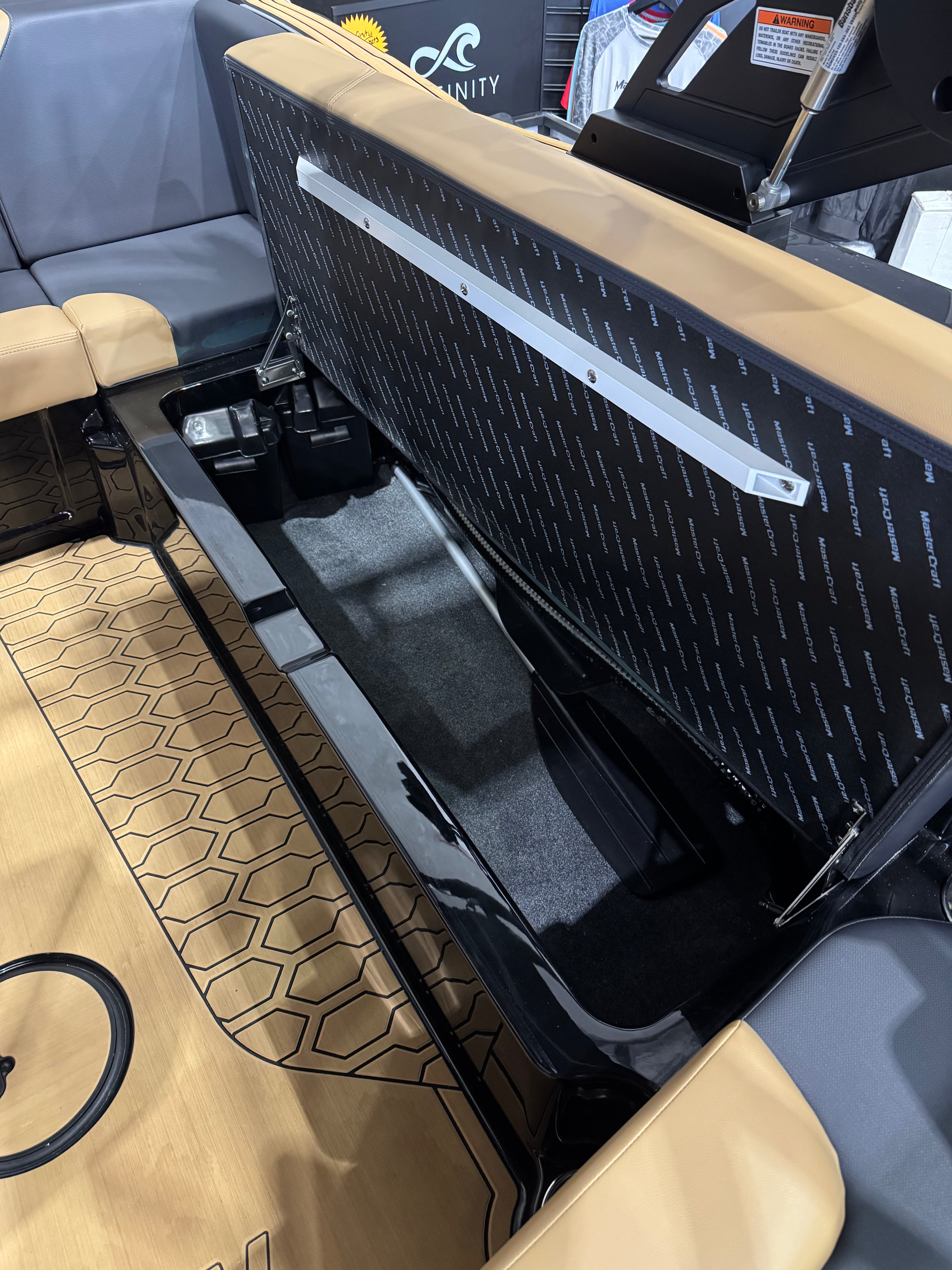 Close-up of a boat interior storage compartment with a black patterned lid, beige seating, and a wood-look floor mat.