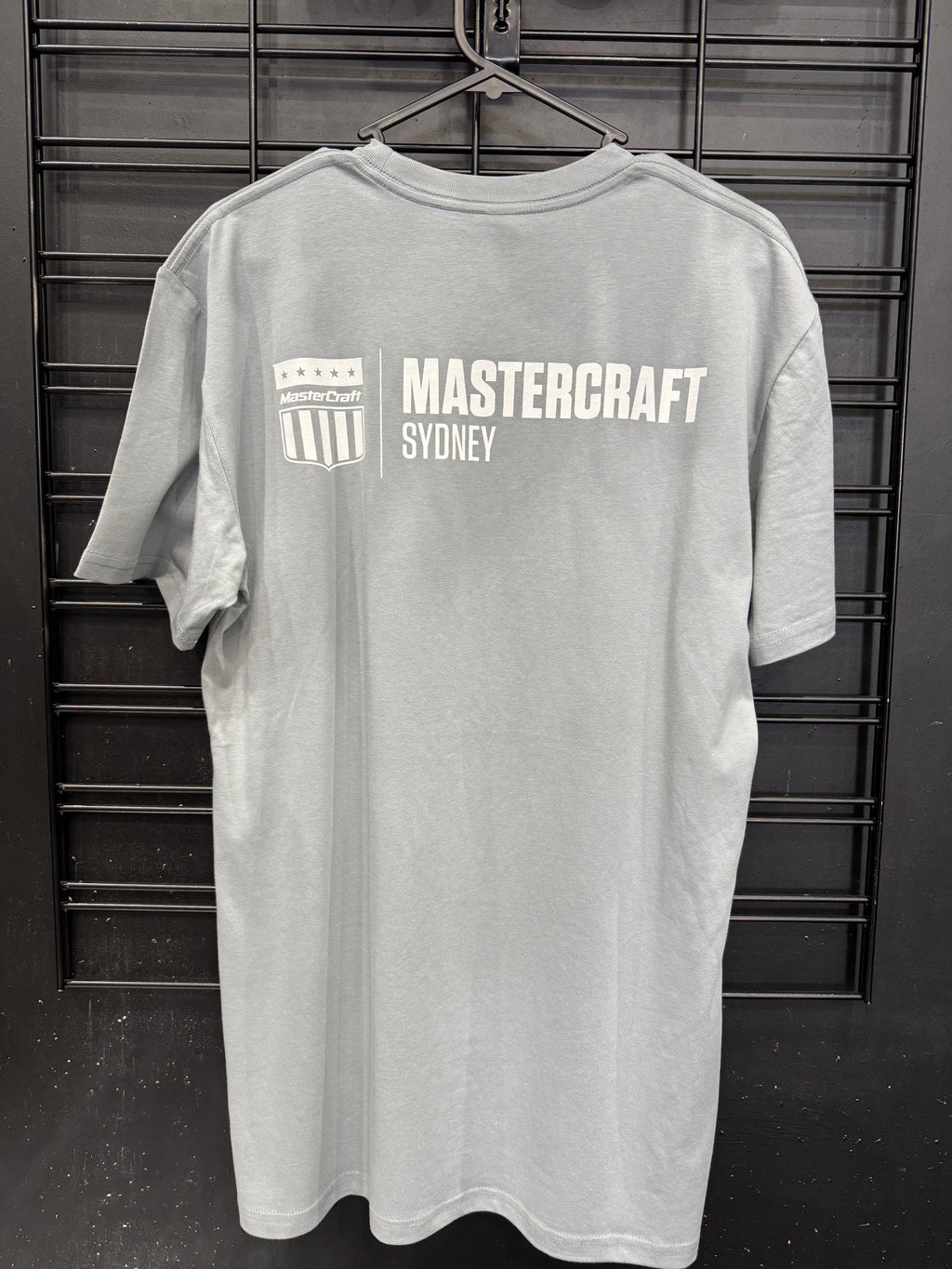 A light gray short-sleeve t-shirt hanging on a black metal hanger against a black slatted wall. The back of the shirt features a white printed graphic with the text 'MASTERCRAFT SYDNEY' and a shield logo.
