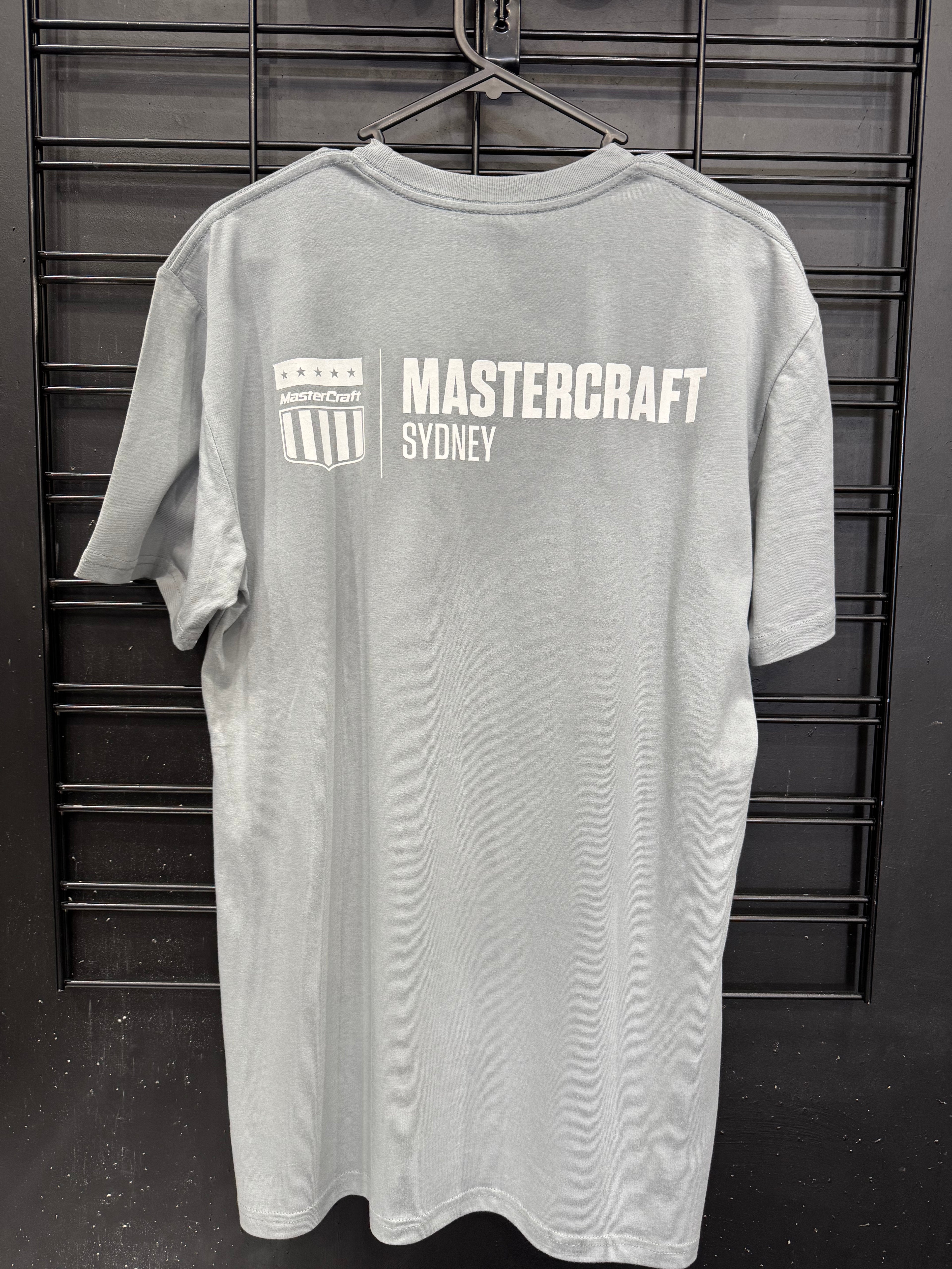 A light gray short-sleeve t-shirt hanging on a black metal hanger against a black slatted wall. The back of the shirt features a white printed graphic with the text 'MASTERCRAFT SYDNEY' and a shield logo.