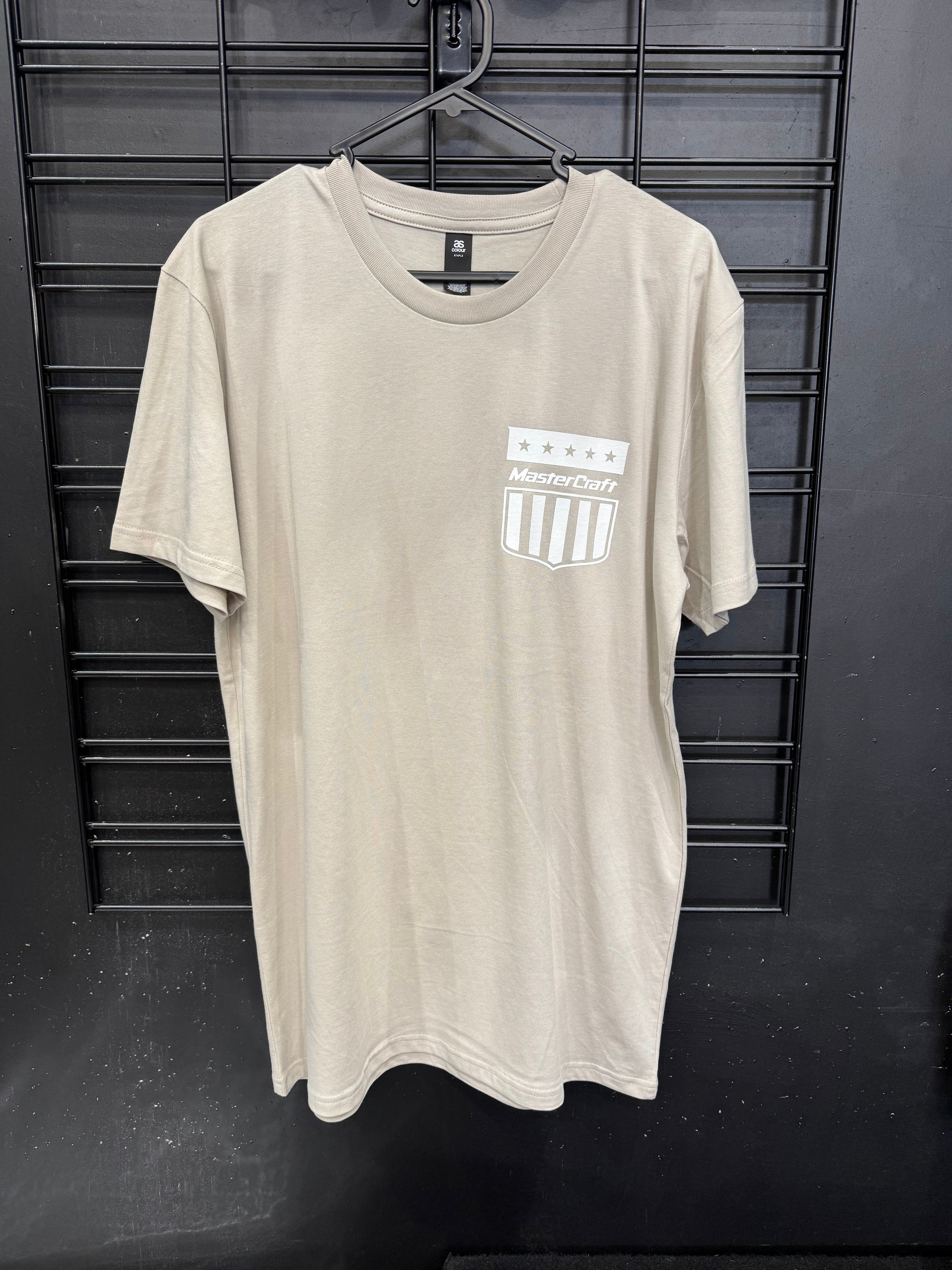 A beige short-sleeve t-shirt hanging on a black hanger against a black metal grid wall. The shirt features a white and gray shield logo on the left chest with the text 'MasterCraft' and five stars above it.