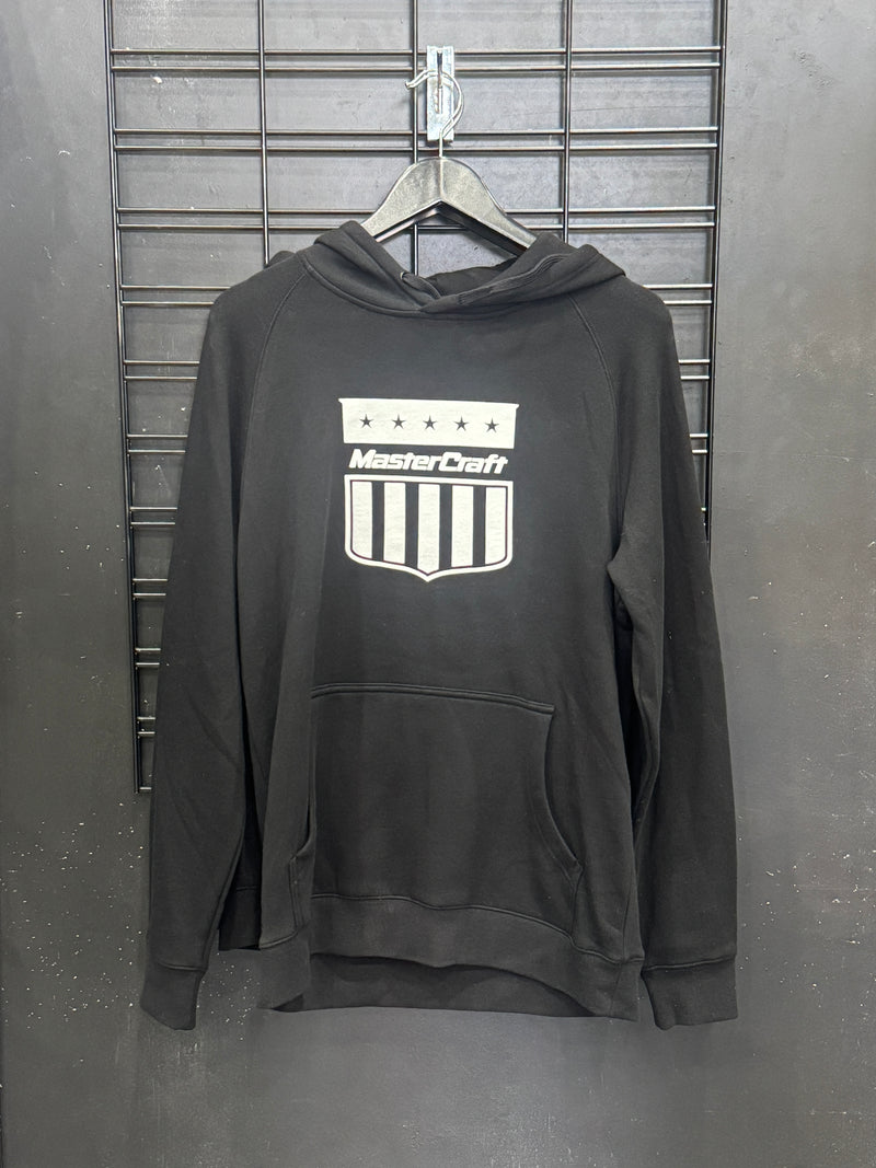 A black pullover hoodie with a white 'MasterCraft' shield logo on the chest, hanging on a black wire hanger against a gray metal grid wall.